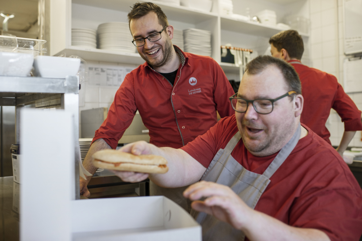 Man pakt broodjes in in doos, andere man kijkt lachend toe