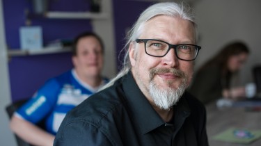 man met bril kijkt lachend in de camera, man en vrouw op de achtergrond