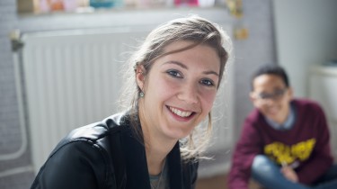 Vrouw kijkt lachend naar de camera, jongen lachend op de achtergrond
