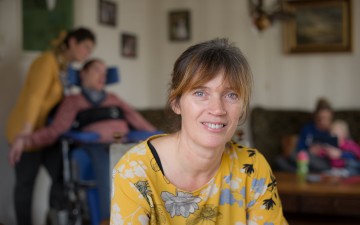 Vrouw kijkt in camera, man in rolstoel op de achtergrond