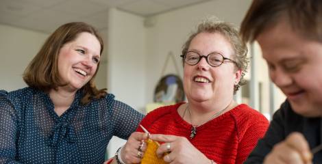 Begeleidster kijkt lachend naar breiende vrouw, andere vrouw op de voorgrond