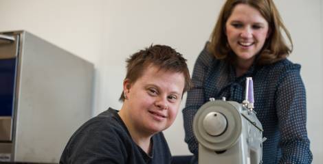 Vrouw achter naaimachine kijkt in de camera, begeleidster staat er naast te kijken